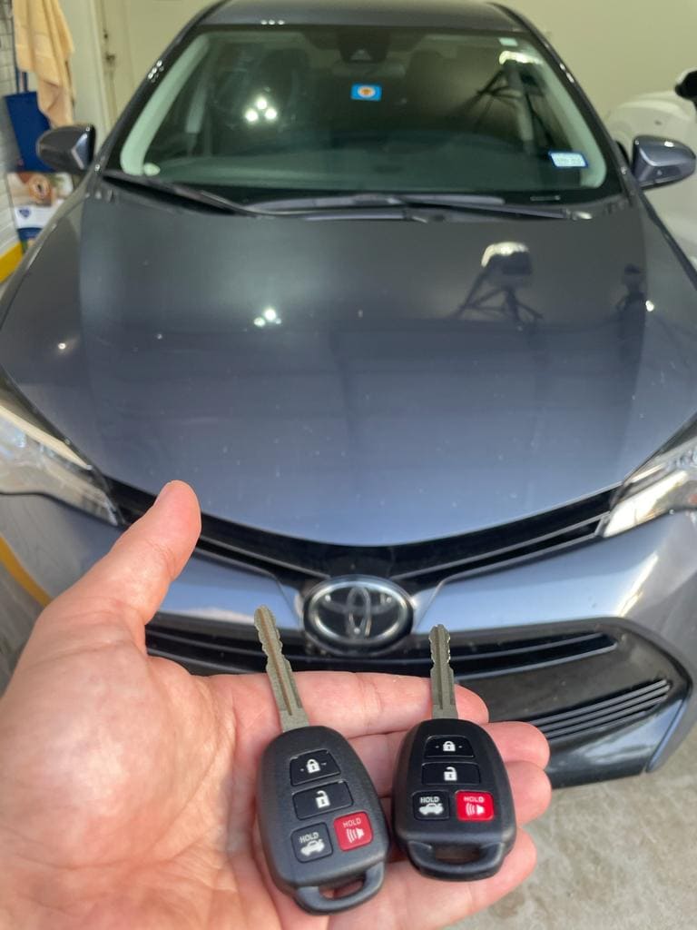 hand holding 2 Lexus car keys next to a blue Toyota car in Houston
