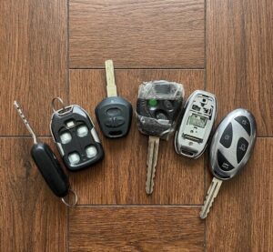 A collection of six different worn and damaged car keys and electronic key fobs arranged in a row on a brown wood tile background, illutsrating common wear and tear issues.