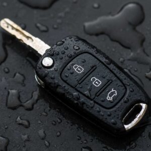 a black electronic car key fob with its physical key blade flipped out, resting on a dark surface covered in water dropless