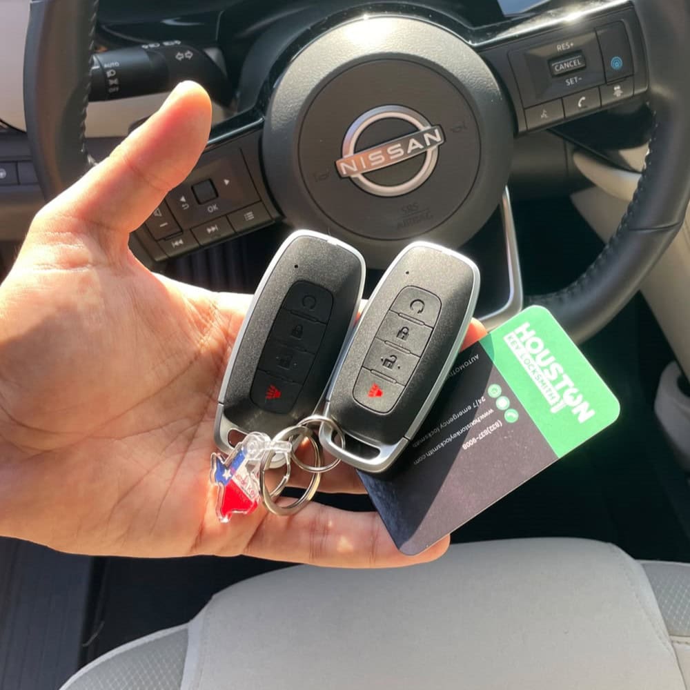 A hand holding two Car Key fobs in front of a steering wheel inside the vehicle with Houston Key Locksmith's Business card